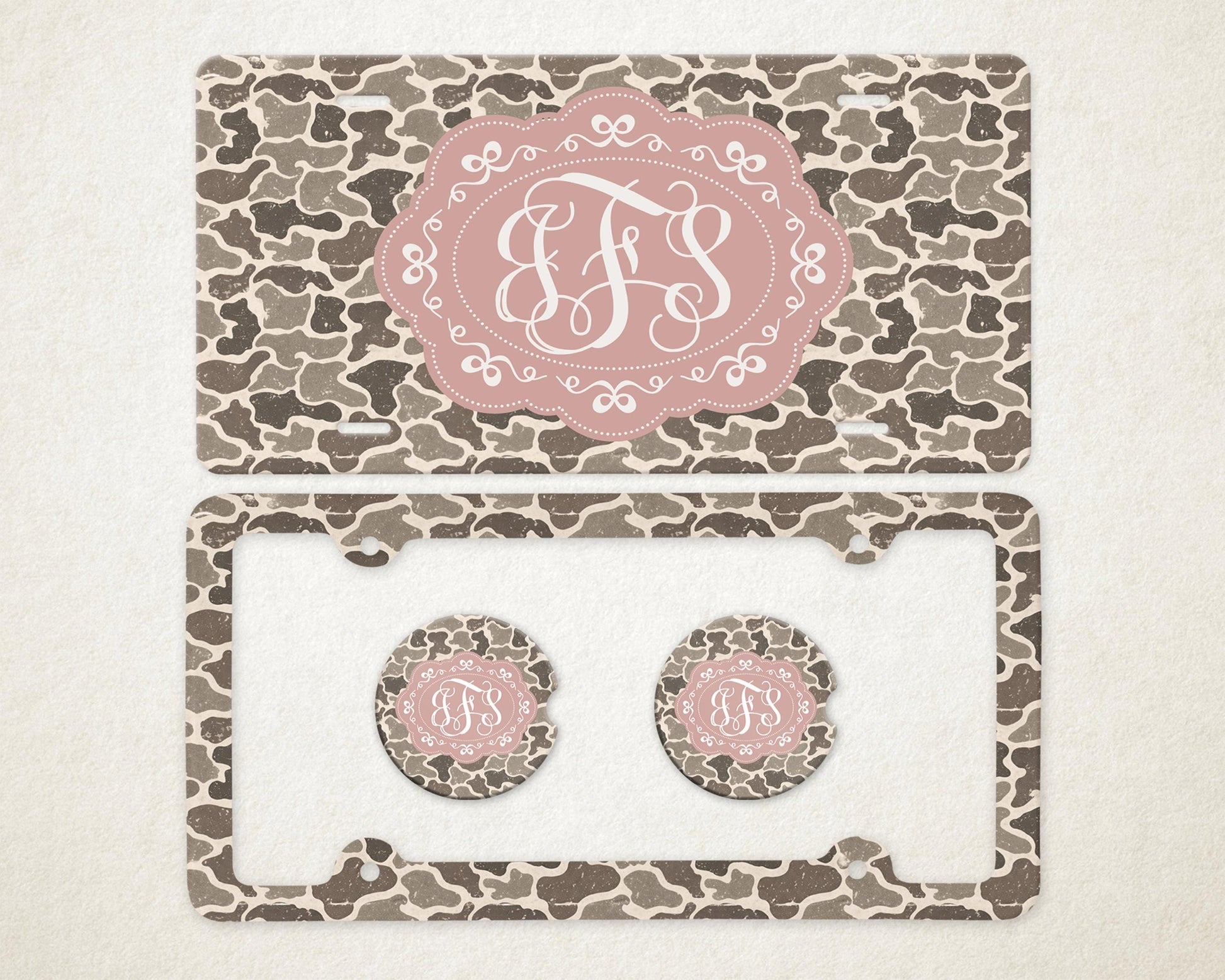 May include: A license plate, license plate frame, and two car coasters with a camouflage pattern in shades of brown and gray. Each item features a pink oval with a white monogram.