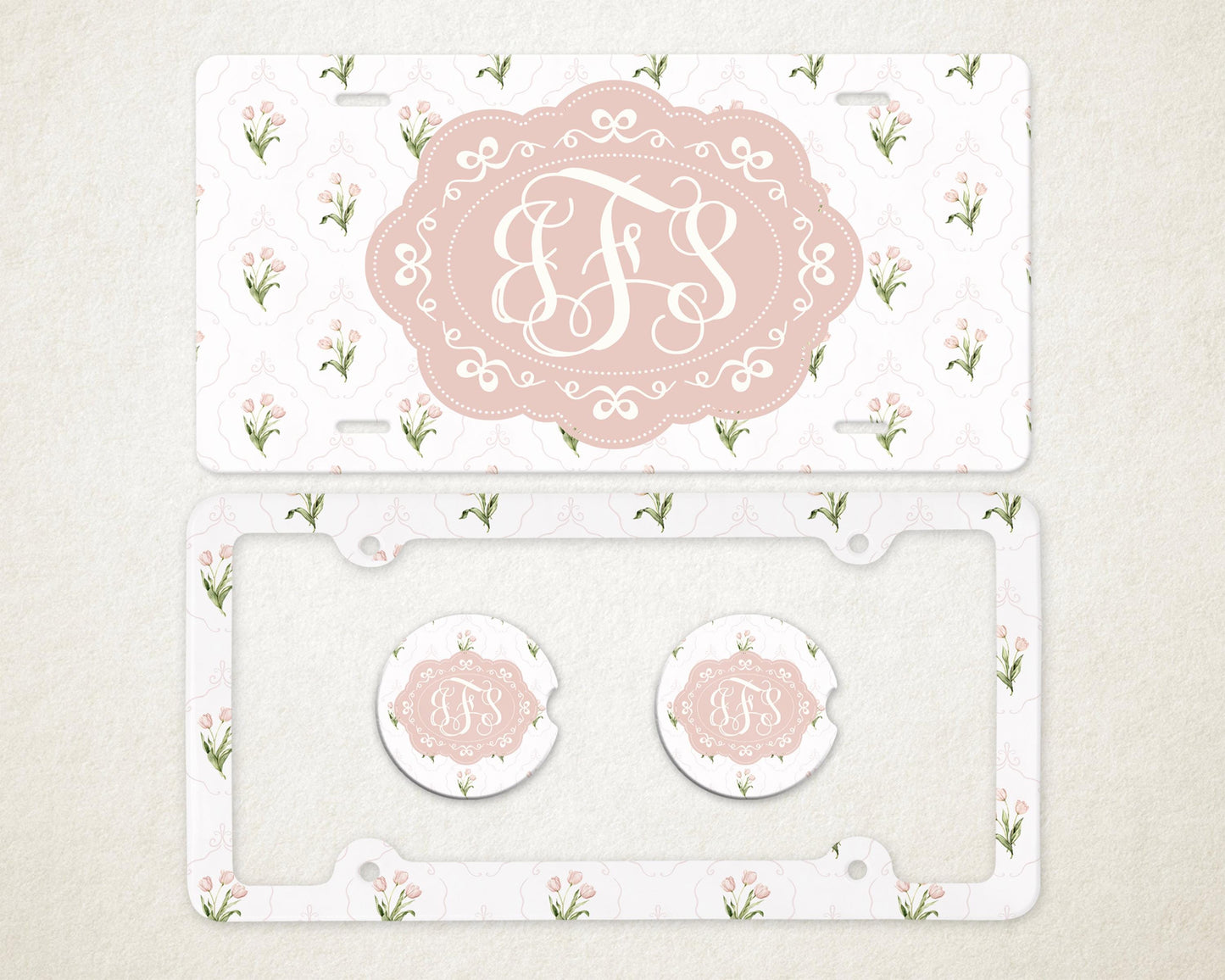 May include: A license plate, license plate frame, and two car coasters with a floral pattern and a monogram. The license plate and coasters have a pink oval with a white monogram. The frame and plate have a white background with pink flowers.